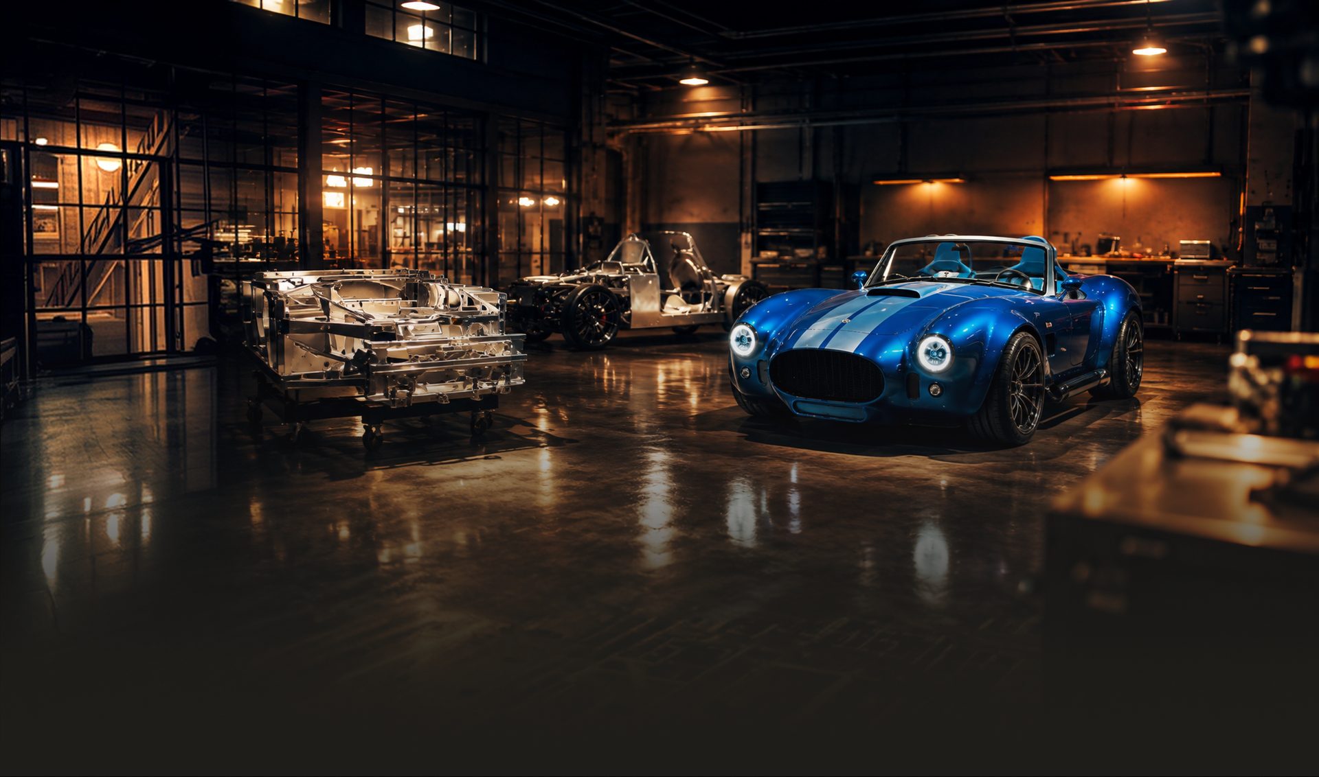 A finished Hinderer roadster in the lit workshop, with a rolling chassis and a bare aluminium tub on its trolley standing beside it.
