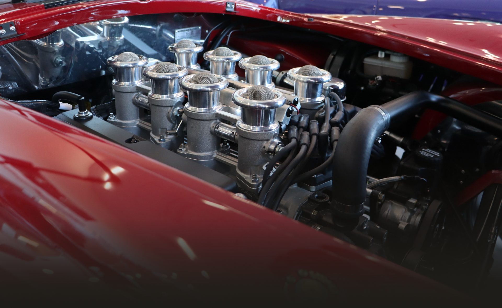 The open bonnet of a red roadster in the workshop, showing eight polished velocity stacks on its injected V8.